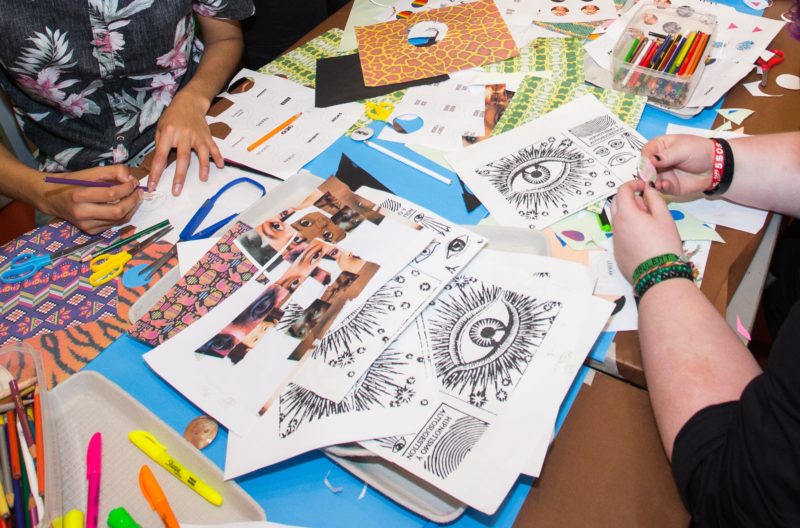 Two sets of hands work on art projects in The Warhol’s studio space. There are colorful pieces of paper on the tables that they are using to collage.