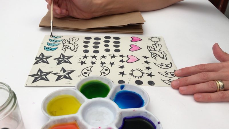 A white palette is in the foreground with orange, yellow, green, and blue watercolor paint. In the background is a pair of hands holding a paintbrush while adding color to white paper filled with stars, moons, hearts and birds stamped in black ink.