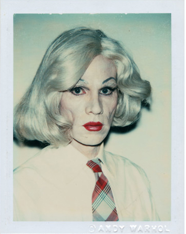 Andy Warhol in drag wearing a white shirt and plaid tie, and medium length silver white wig. He has white pancake make-up covering heavily covering his face, red lips, and heavily made-up eyes. He is looking at the camera in three-quarter profile, not smiling. His name is embossed on the photograph in the lower right-hand corner.