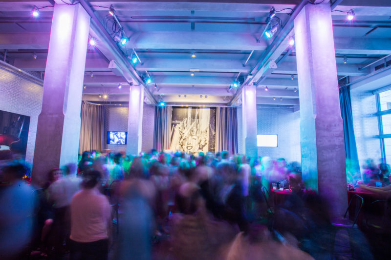 A group of people dancing and sitting at tables. On the wall behind the group, a picture of Andy Warhol holding up a large acetate of Marilyn Monroe’s face is visible.