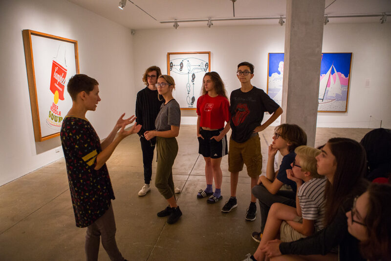 A Warhol educator stands to the left, speaking to an audience and gesturing with her hands. Bright, large, colorful Andy Warhol paintings hang on the walls of a gallery space in the background. Her audience is mostly teenagers. Some are standing in the middle ground behind her, and most are seated to the right, watching her intently.