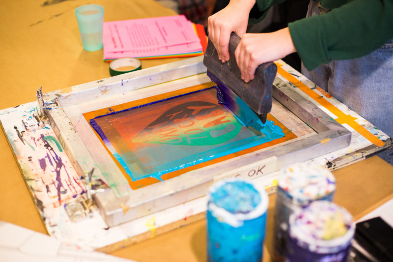 Two hands on a squeegee, pulling blue and purple ink over a silkscreen image of a teardrop with the words “it’s ok to cry baby” inside.