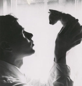 A black and white photograph of Andy Warhol holding a kitten in front of a window draped in white cloth.