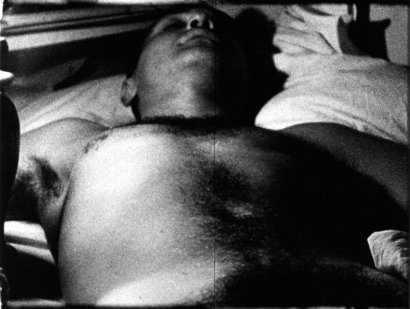 A still from one of Warhol's black and white films featuring a shirtless man lying on his back, asleep.