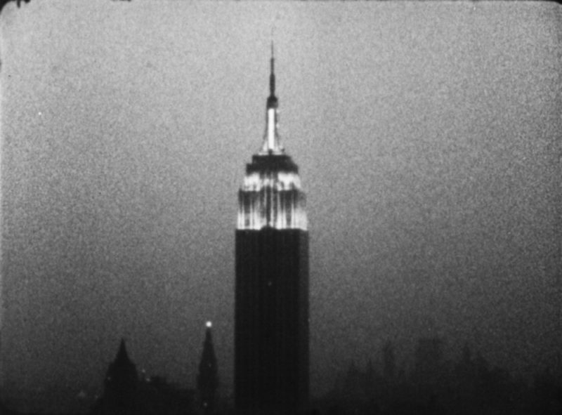 A grainy black and white image of the top of the Empire State building, illuminated in the dusk.