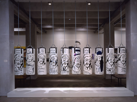 Ten punching bags which have been painted white with black words and line-drawings of Jesus's face hang in a row by silver chains attached to a high ceiling.
