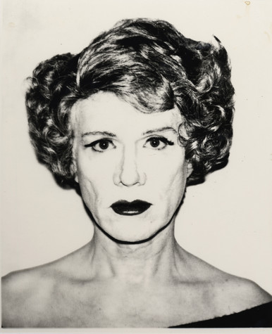 A photograph of Andy Warhol dressed in drag. He wears very pale face makeup, a bold lip, and a short, curly wig.