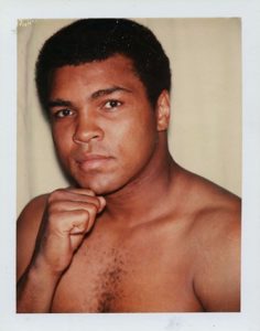 A photograph of boxer Muhammed Ali shows Ali standing against a pale background and poses for the camera, his right fist raised to his chin.