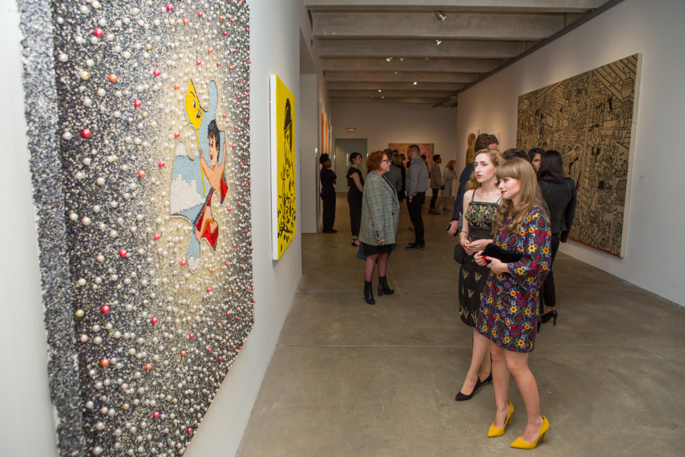 Two women look at a canvas covered in an intricate design made with beads of various colors and sizes.