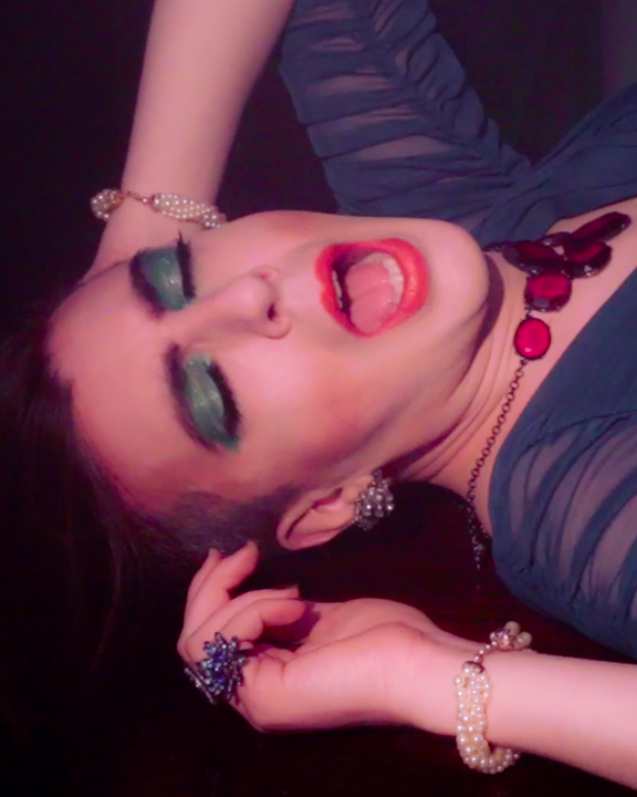 The image features artist Atom Atkinson. They are wearing a sheer, turquoise top, big ruby-colored necklace, braided pearl bracelet on both wrists, and blue flower ring. They also have on sparkly blue eyeshadow and coral lipstick. Their mouth is open, as if they are singing.