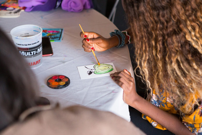 A person’s hands are visible completing a watercolor painting at a table under an outdoor tent.