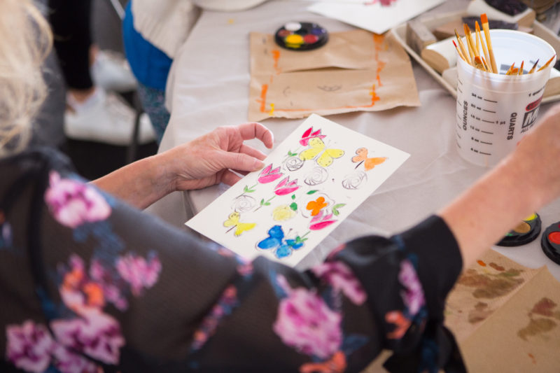 A person is sitting at a table with art supplies holding a partly painted coloring page with outlines of butterflies and flowers on it.