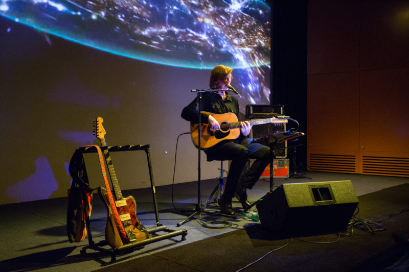 A man with a guitar sits on a dark stage with a projection of outer space on the wall behind him.