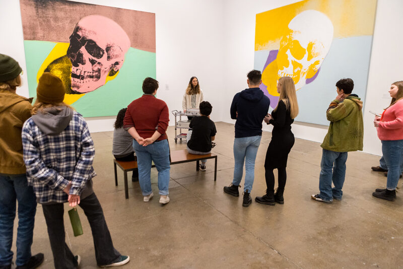 A group of people are in a gallery in The Andy Warhol Museum with two large screenprints of Warhol's Skull series hanging on the walls. The people are facing one person who appears to be giving a tour.