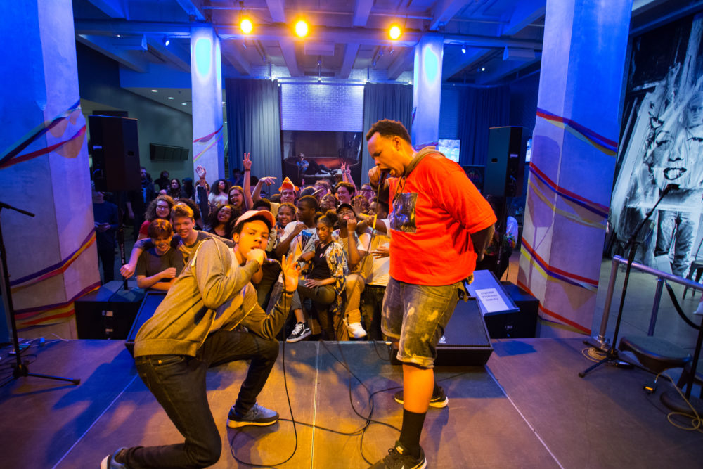 A man in a gray hoodie and baseball cap and a man in a red t shirt sing into microphones as a crowd of teens in the Andy Warhol Museum entrance space cheer behind them.