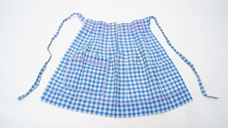 Front view of a blue gingham apron. In addition to the gingham pattern, the apron has pink cross-stitched accents.