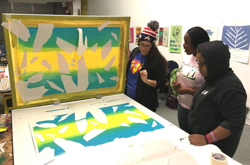Three women, members of the 2018 Youth Arts Council, are standing off the the right of the frame, looking at a screenprint they have just created at Artist Image Resource. The woman on the left is holding up the screen.
