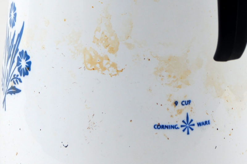 View of the coffee pot’s back side showing the CorningWare logo and a blue floral design. There is a black handle with a metal top.