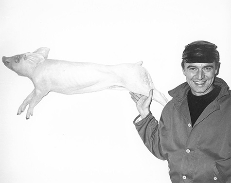 Artist Rene Ricard stands next to an art piece depicting a pig on its side.