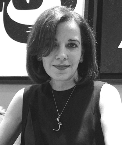 The photo is a black and white portrait of a woman with dark hair and eyes. Her hair reaches her shoulders and is parted on the side, a portion covers her left eye. She is looking directly at the camera and smiling. She is wearing a dress that reveals her shoulders and has accessorized it with circular earrings and a necklace with a charm at the end, it appears to be a symbol or letter. Behind her one can see a portion of two abstract paintings, very much with patterns similar to her necklace.