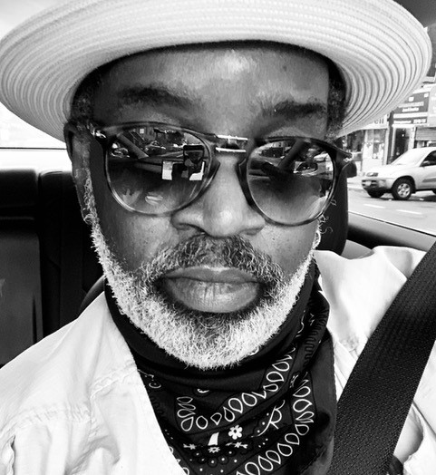 Black and white photograph of a person with a beard and mustache in a car with a seatbelt on looking towards the viewer, wearing a light-colored hat, sunglasses, white shirt, and a dark bandana around their neck.