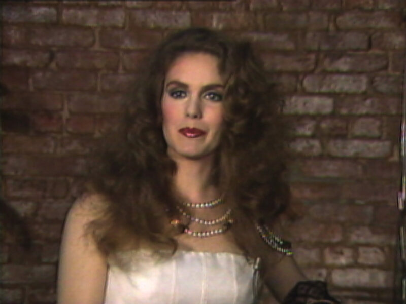A video still of a person in front of a red brick wall with long, wavy brown hair wearing blue eye shadow, pink blush, and red lipstick, three pearl necklaces, and a white dress.