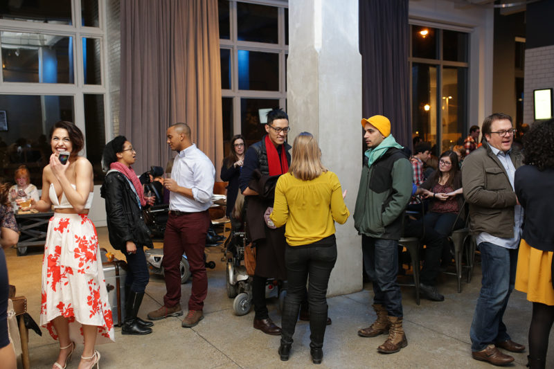 An exhibition opening event taken in The Andy Warhol Museum entrance space. It is crowded with people, who are talking in pairs and small groups in the foreground with beverages in hand. On the left side of the image, a woman in a white sundress with an orange floral print is laughing, holding her left hand to her mouth.