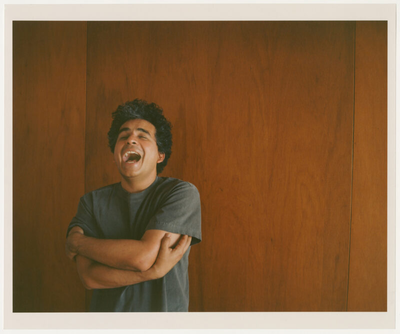 Helado Negro stands in front of a wood wall with arms crossed and his mouth open wide.