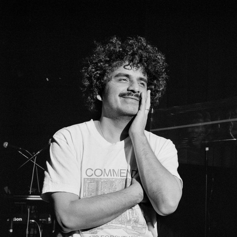 Man with full head of curly black hair and a mustache crosses his arms and looks off into the distance.