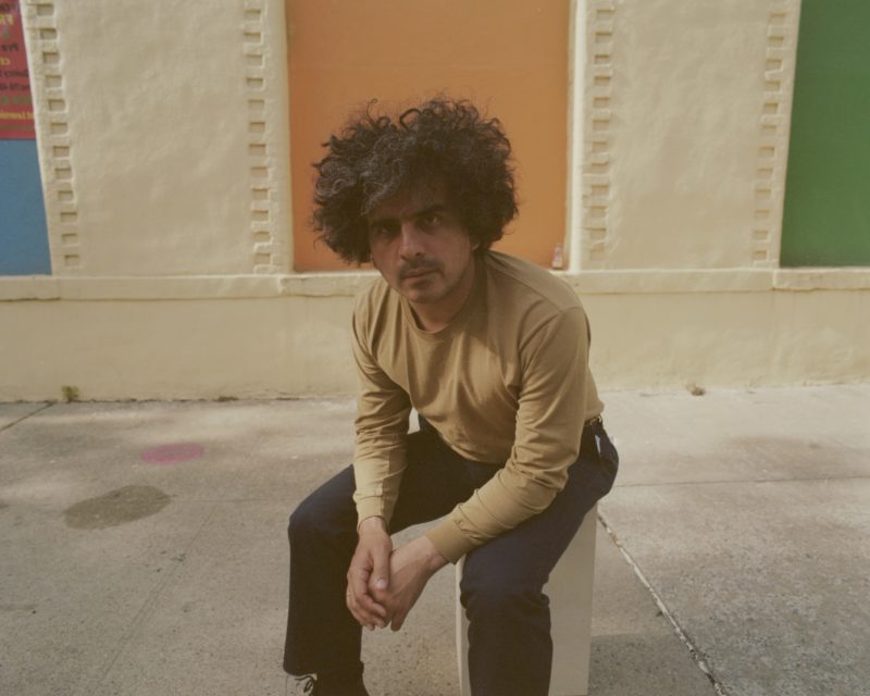 A person with dark hair, wearing a tan shirt and blue pants, sits on a block looking towards the camera.