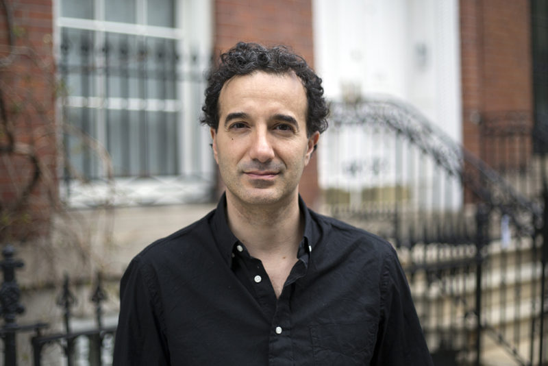 A person from the chest up wearing a black button down shirt taken in front of a brick building.