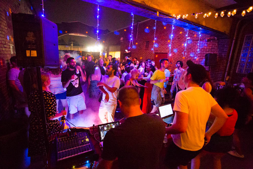 A group of three DJ's are touching laptops and DJ equipment while a crowd dances in front of them.