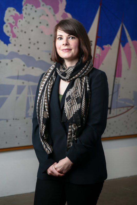 A person who is smiling stands in a gallery in front of an artwork by Andy Warhol.