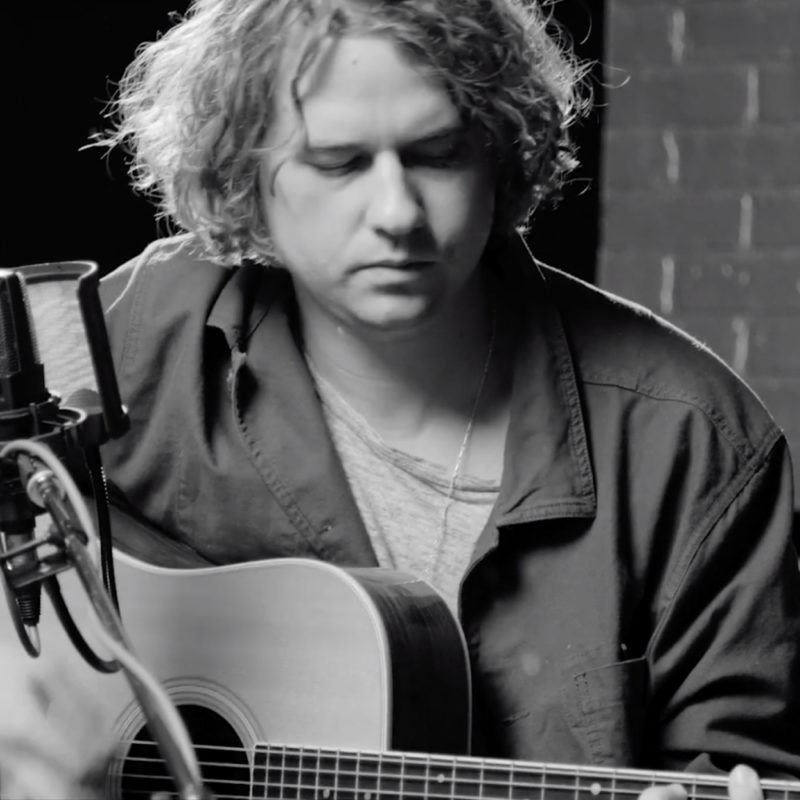 Man with guitar sits behind a microphone.