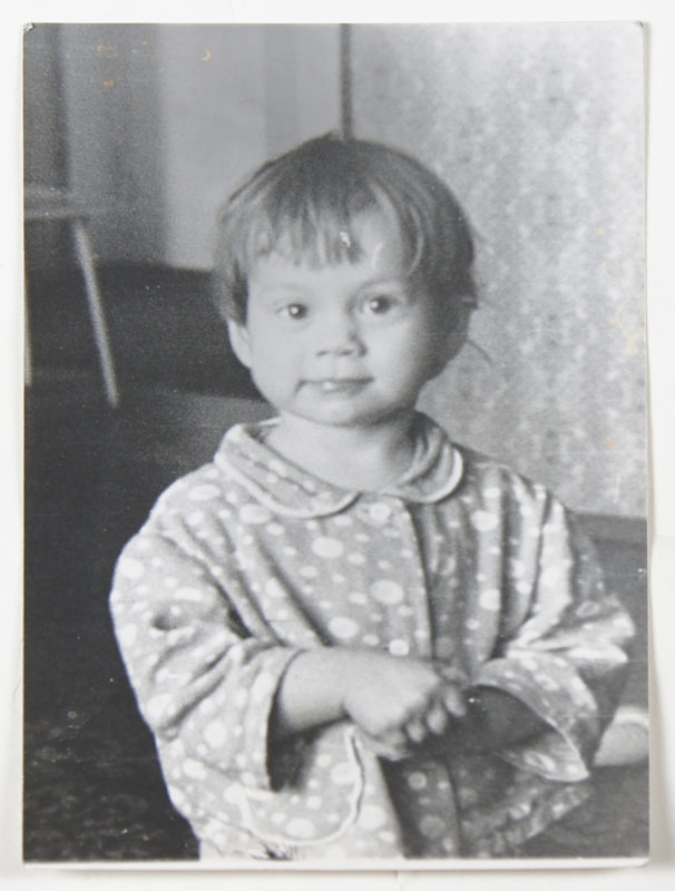 Black and white photo of a toddler with short hair wearing a flowered top and with her hands clasped in front of her.