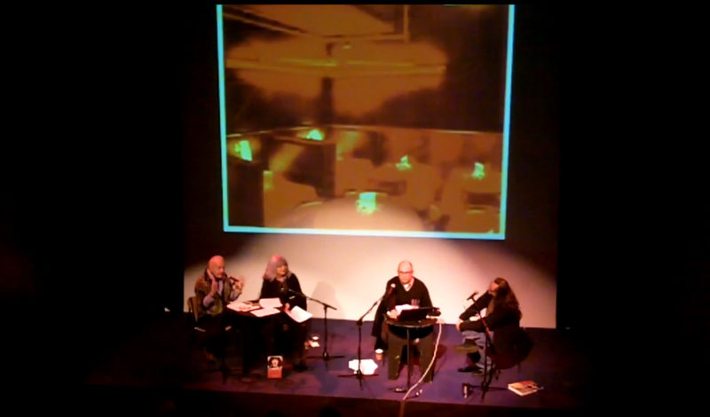 Four people sit on a stage with two tables and a three microphones.