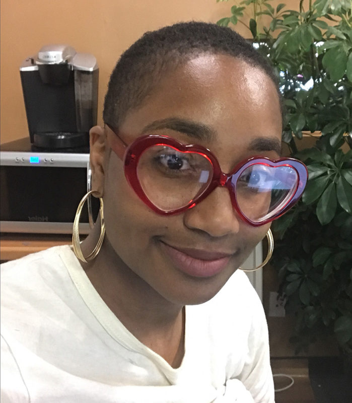 A person wearing red, heart-shapped glasses and a white shirt smiles for the camera