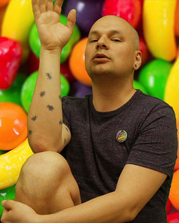 A bald man is pictured in the center of the image. His right knee is bent and positioned at the foreground. His right elbow rests on his knee, as he lifts his hand up, with his palm facing outward. His forearm is covered in spider tattoos. An image of bright, fruit-shaped candy forms the background of the image.