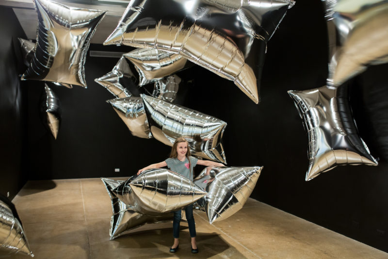 A young girl with long brown hair wearing a black and white striped shirt and skinny jeans interacts with large, rectangular balloons in The Andy Warhol museum’s Silver Clouds exhibition.