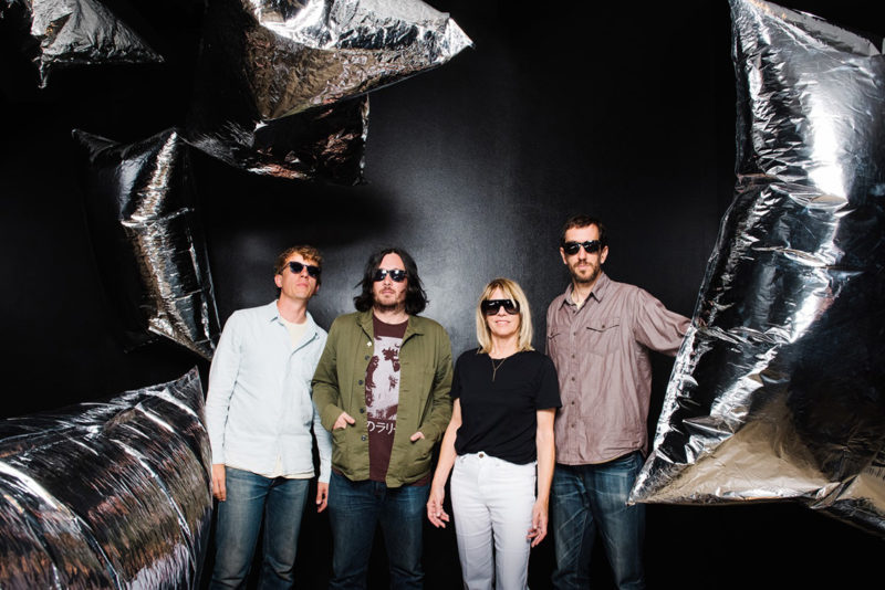 Three men and one woman stand in a gallery with black walls as silver balloons float around them.