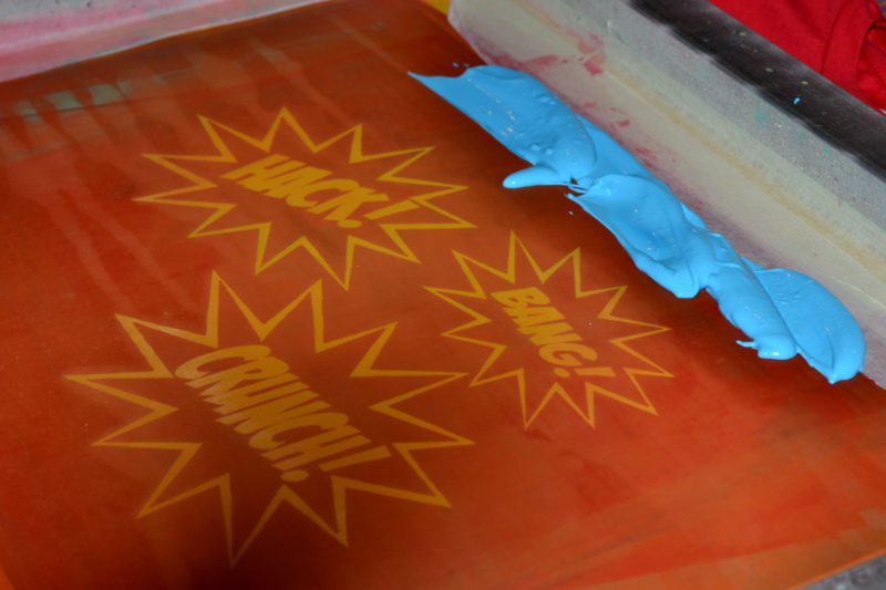 A silkscreen with the words hack, bang, and crunch designed with a Pop art treatment. Blue paint is on the top of the screen, waiting to be pulled across it.