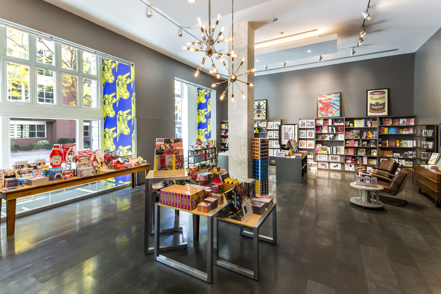 A photograph of the Warhol store. The far wall is occupied by book shelves, and there is a table full of products pushed against the windows overlooking the street on the left side of the photo. Another display occupies the center of the image, under three hanging wheel-like light fixtures.