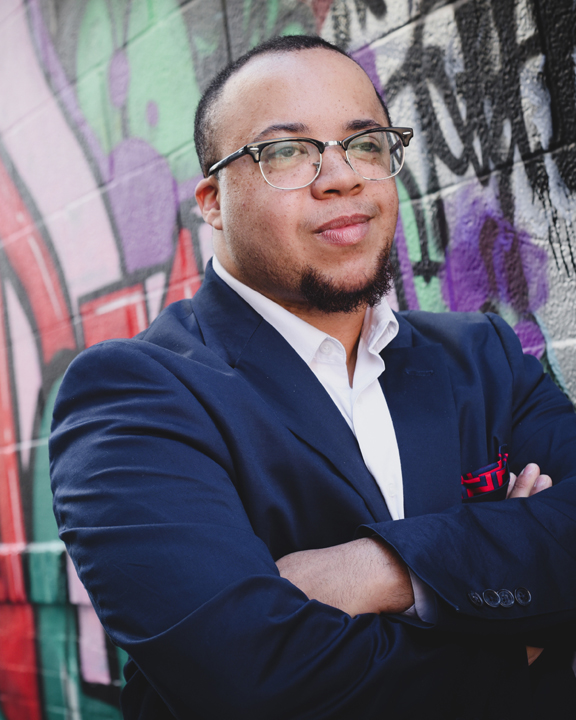 A man stands with his arms crossed at the foreground of the image. He is wearing glasses, a white button-down shirt, and navy blazer. A brick wall painted with brightly colored graffiti makes up the background.