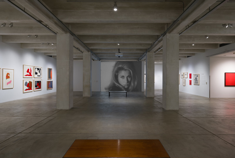 Installation view of a gallery with several framed prints and paintings and one large (center) projection of a black and white film portraying a woman looking directly into the camera.
