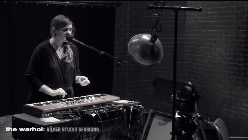 A long-haired person standing at a keyboard speaks into a microphone.