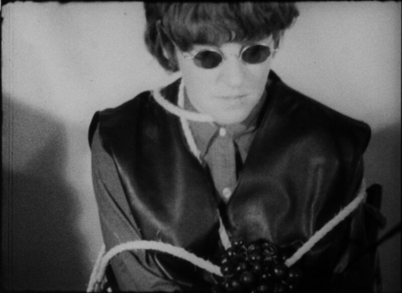 Black and white film still of a person with short, dark hair, sitting down and wrapped with a rope.