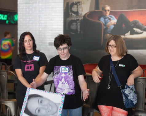 The boy in the center of the image tosses a large cube with a picture of a smiling young women on the viable side of the cube. His mother, to his left, looks on smiling.