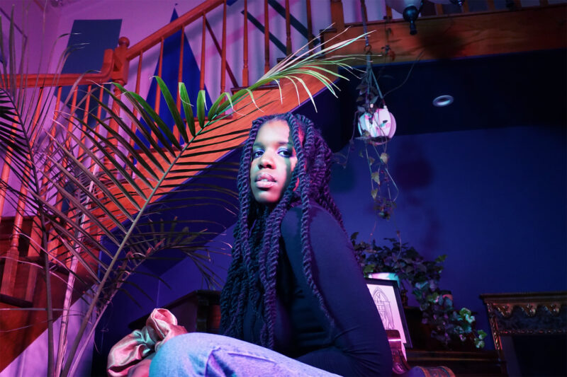 Zenizen sits on a chair in front of a staircase while looking down towards the camera.